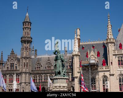 Eine Statue vor herrlichen gotischen Gebäuden mit wehenden Fahnen unter einem klaren blauen Himmel, Brügge, Belgien Stockfoto