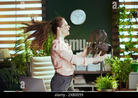 Nachhaltiger Arbeitsplatz. Lächelnde, moderne 40-jährige Angestellte im modernen grünen Büro mit elektrischem Ventilator. Stockfoto