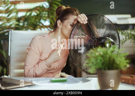 Nachhaltiger Arbeitsplatz. Moderne Arbeitnehmerin mit elektrischem Ventilator, der unter Sommerwärme leidet. Stockfoto