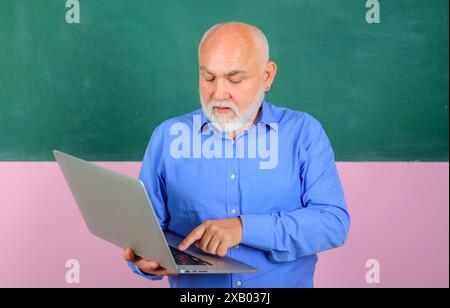 Männlicher Lehrer in blauem Hemd mit Laptop-pc im Klassenzimmer. Bildung in der High School oder am College. Ernsthafter Lehrer in der Nähe der Tafel, der le gibt Stockfoto