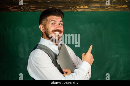 Lächelnder bärtiger Lehrer mit Laptop-Computer, der mit dem Finger auf die Tafel zeigt. Männlicher Lehrer, der den Schülern Unterricht gibt. Ausbildung bei hoher sch Stockfoto