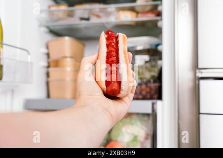 Nahaufnahme einer Hand nimmt einen Hot Dog aus dem Kühlschrank mit Ketchup vor einem offenen Kühlschrank mit Lebensmittelbehältern und Lebensmitteln. Stockfoto