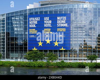 Straßburg, Frankreich - 6. Juni 2024: Nahaufnahme des Gebäudes des Europäischen Parlaments in Straßburg mit einem großen Banner in mehreren Sprachen, das die Wahl bei den EU-Wahlen 2024 anregt, mit Reflexionen auf der Glasfassade und einem Fluss im Vordergrund. Stockfoto