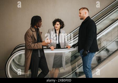 Drei Geschäftsleute in Winterkleidung führen ein freundliches Gespräch, während sie eine Rolltreppe in einem modernen Geschäftsumfeld hinabsteigen. Stockfoto