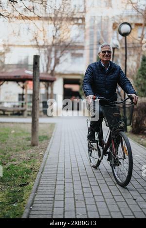 Ein aktiver Seniorenmann mit Brille fährt mit dem Fahrrad durch einen ruhigen Stadtpark, der Vitalität und einen fröhlichen Lebensstil ausstrahlt. Stockfoto