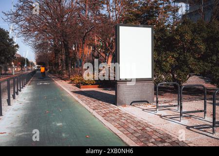 Ein leeres Reklametafelmodell steht neben einem Fahrradweg auf einem von Bäumen gesäumten Weg in einem Stadtpark. Eine Silhouette eines Radfahrers mit einer orangefarbenen Jacke fährt aw Stockfoto