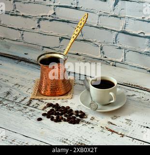 Kupfertürke, geröstete Kaffeebohnen und eine Tasse frisch gebrühten schwarzen Kaffee auf einer Untertasse mit einem Löffel auf einem hellen Holztisch. Nahaufnahme. Stockfoto