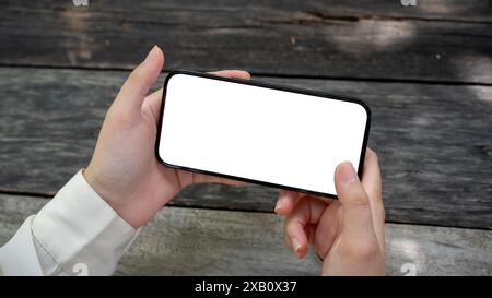 Ein Nahbild einer Hand einer Frau, die ein Smartphone mit einem White-Screen-Modell in horizontaler Position über einem Holztisch im Garten hält. Personen und Stockfoto