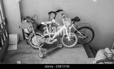 Cremona, Italien - 16. Mai 2024 drei Kinderfahrräder und ein Roller stehen auf einer Veranda mit einem Mülleimer in Schwarz-weiß Stockfoto