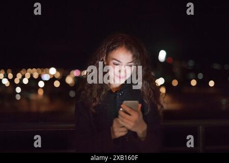 Schöne Frau, die nachts auf der Terrasse steht, telefoniert, Smartphone hält, scrollt. Stockfoto