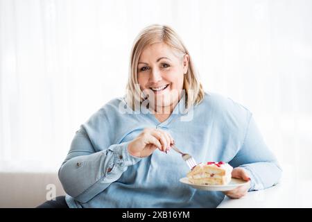 Porträt einer schönen jungen übergewichtigen Frau zu Hause, die ein Stück Kuchen isst, süßen Geschmack genießt. Stockfoto