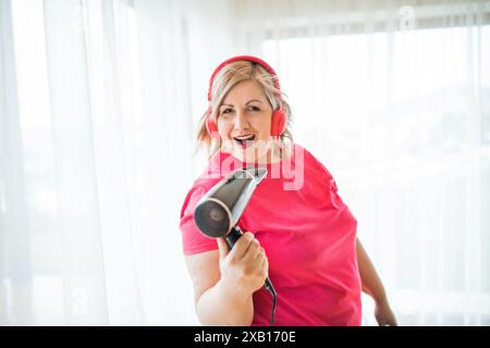 Porträt einer schönen jungen übergewichtigen Frau zu Hause, Musik über Kopfhörer hören, Spaß haben, in den Haartrockner singen. Stockfoto