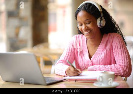 Glücklicher schwarzer Schüler, der Notizen macht, E-Learning mit Laptop und Kopfhörern in einer Bar Stockfoto