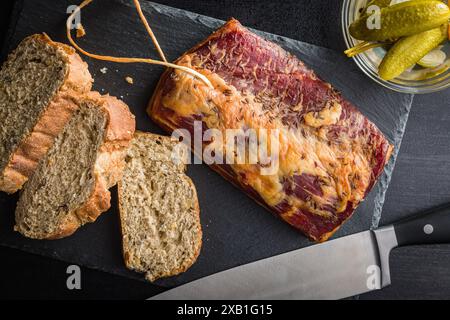 Ein Nahbild eines aufgeschnittenen geräucherten Schweinebauchs, serviert mit Brot und Gurken. Der Schweinebauch wird mit Kümmel abgeschmeckt und auf einem schwarzen Schiefer gelegt Stockfoto