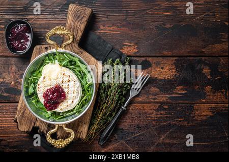 Gebackener Camembert Brie-Käse mit Preiselbeersauce und garniert mit Rucola-Salat in einer Pfanne. Holzhintergrund. Draufsicht. Speicherplatz kopieren. Stockfoto