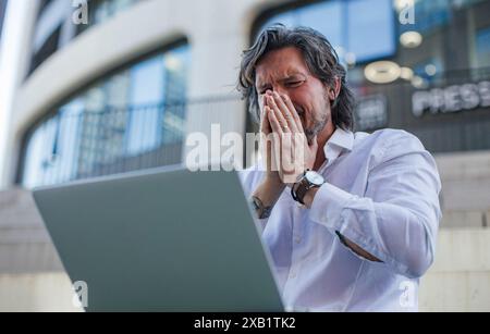 Porträt eines Reifen Geschäftsmannes, der an einem Laptop im Freien arbeitet und schlechte Nachrichten erhält. Schöner Mann mit Brille im Anzug. Stockfoto
