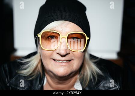 Eine ältere Frau trägt schwarze Mütze und gelbe Brille und lächelt drinnen Stockfoto