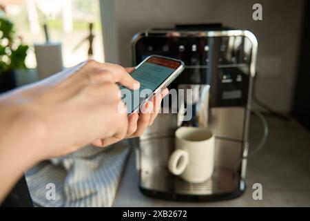 Die Person verwendet die Smartphone-App, um die Kaffeemaschine zu bedienen. Frau, die eine frische Tasse Kaffee mit moderner Espressomaschine zubereitete Stockfoto