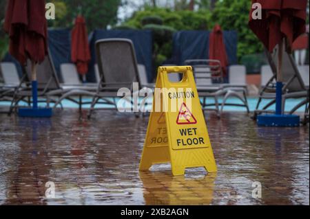 Gelbes Warnschild mit der Aufschrift „Caution Wet Floor“, neben dem Swimmingpool und den Sonnenliegen eines Hotelresorts. Stockfoto