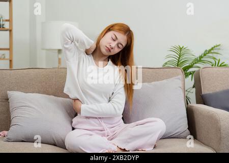 Junge Frau, die sich zu Hause unwohl fühlt, mit Rückenschmerzen auf der Couch sitzt, bequeme Loungewear trägt, in einem modernen Wohnzimmer mit Soft Stockfoto