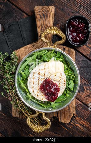 Gebackener Camembert Brie-Käse mit Preiselbeersauce und garniert mit Rucola-Salat in einer Pfanne. Holzhintergrund. Draufsicht. Stockfoto