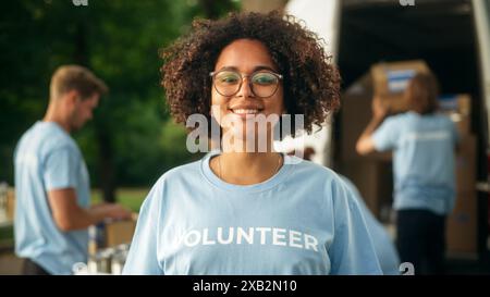 Porträt einer glücklichen hilfsbereiten Schwarzen Freiwilligen. Junge Erwachsene multiethnische Latina mit Afro-Haar, Brille, Lächeln, posieren für die Kamera. Konzept für humanitäre Hilfe und Freiwilligentätigkeit. Stockfoto