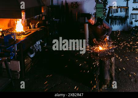 Ein männlicher Schmied arbeitet aufmerksam und hämmert ein Stück heißes Metall auf einen Amboss, umgeben von der Glut und den Funken in einer schwach beleuchteten Schmiede. Stockfoto