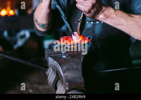 Ein abgeschnittener, nicht erkennbarer männlicher Schmied schmiedet eine Metallblume auf einem Amboss in einer traditionellen Werkstatt mit Feuerhintergrund. Stockfoto