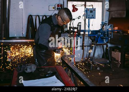 Schmied arbeitet mit einem Winkelschleifer an einer Stahlkonstruktion und erzeugt Funken in einer Werkstatt. Stockfoto