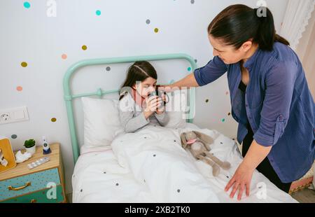 Eine fürsorgliche Mutter hilft ihrer kranken Tochter, aus einer Tasse zu trinken, während sie in einem gemütlichen Schlafzimmer sitzt, umgeben von Komfortgegenständen Stockfoto