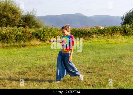 Eine fröhliche junge Frau in farbenfroher Kleidung tanzt auf einer Wiese mit der malerischen Magina Mountain Range im Hintergrund. Stockfoto
