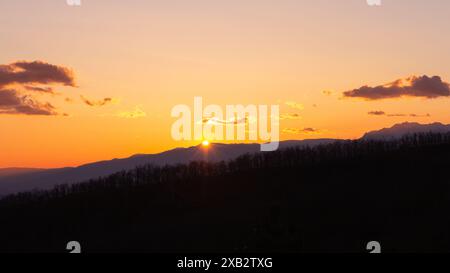 Eine atemberaubende Szene des Sonnenuntergangs hinter einer Bergkette, die ein warmes Leuchten über den Himmel wirft und Silhouetten der Gipfel dieses friedlichen, swee hervorhebt Stockfoto