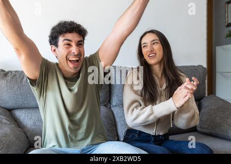 Ein fröhliches junges Paar genießt einen Moment des Sieges, während er zu Hause auf einem Sofa sitzt. Der Mann hebt aufgeregt die Arme, während die Frau zusieht und smi Stockfoto