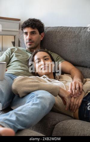 Ein liebevolles Paar liegt bequem auf einem Sofa und teilt einen intimen Moment der Entspannung. Der Mann hält einen Becher, während die Frau sanft ihren Kopf auf h legt Stockfoto