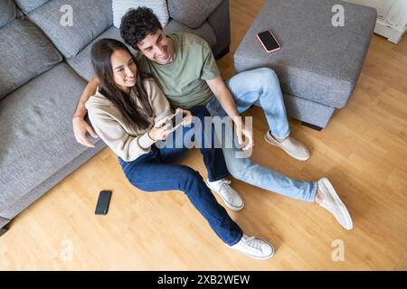 Ein junges Paar sitzt auf dem Boden in ihrem modernen Wohnzimmer und teilt einen fröhlichen Moment, während es gemeinsam Videospiele spielt, und zeigt eine Mischung aus Spaß und Spaß Stockfoto