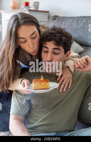 Ein junges Paar genießt einen gemütlichen Moment zu Hause, während die Frau dem Mann eine Geburtstagskuche mit einer zündeten Kerze überreicht und zeigt, dass sie den Kerzenständer ausblasen Stockfoto