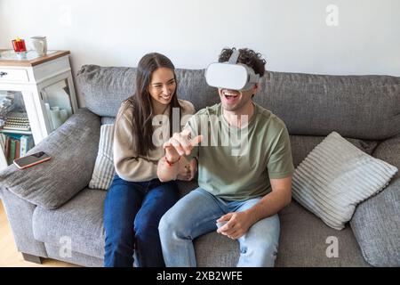 Eine junge Frau lacht neben einem Mann, der ein VR-Headset trägt, und spielt spielerisch in einem modernen Wohnzimmer Stockfoto