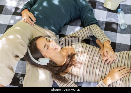 Ein junges Paar genießt einen ruhigen Moment zusammen im Freien, liegt auf einer karierten Picknickdecke. Die Frau hört Musik über Kopfhörer, während sie sich ausruht Stockfoto