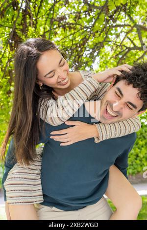 Ein junges Paar teilt einen freudigen Moment im Freien, mit dem Mann, der die Frau auf dem Rücken trägt, beide lachen unter einem Baldachin aus üppig grünen Blättern Stockfoto