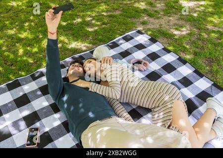 Ein junges Paar liegt auf einer karierten Decke in einem üppigen Park und hält einen Selfie-Moment im Schatten der Bäume fest, umgeben von Grün und Picknick essen Stockfoto
