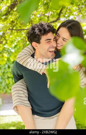 Ein fröhliches junges Paar teilt einen verspielten Moment inmitten des lebendigen Gartens, während der Mann der Frau einen Huckepack-Ritt gibt Stockfoto