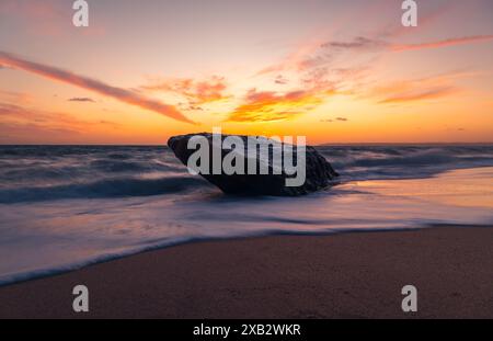 Leuchtende Sonnenuntergänge prägen den Himmel über Atlanterra Beach, während die Wellen einen einsamen Felsbrocken am Sandstrand streicheln. Stockfoto