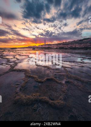 Ein atemberaubender Sonnenuntergang über felsigen Gezeitenbecken mit dem Trafalgar Lighthouse im Hintergrund in Cadiz. Stockfoto