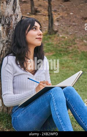 Eine besinnliche Frau sitzt an einem Baum in einer ruhigen Parklandschaft und schreibt nachdenklich in ihr Notizbuch, um den Frieden der Natur zu genießen Stockfoto