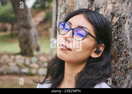 Eine ruhige junge Frau lehnt sich mit geschlossenen Augen an einen Baum und genießt einen friedlichen Moment in einer üppigen Parklandschaft Stockfoto