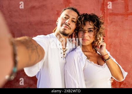 Ein junges Paar teilt einen Moment der Verbindung, während es ein Selfie vor einem leuchtend roten Hintergrund macht, die Frau verführt den Blick und den Mann, der ein Lächeln begrüßt Stockfoto
