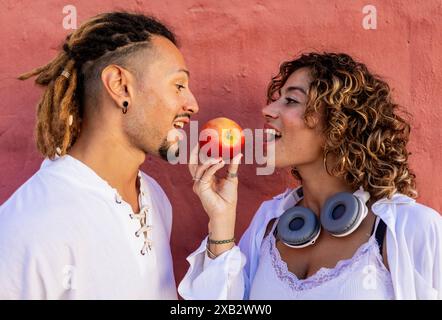 Ein junger Mann und eine junge Frau teilen sich einen roten Apfel und lächeln sich verspielt an einer leuchtend roten Wand an. Die Frau trägt weiße Kopfhörer um den Hals Stockfoto