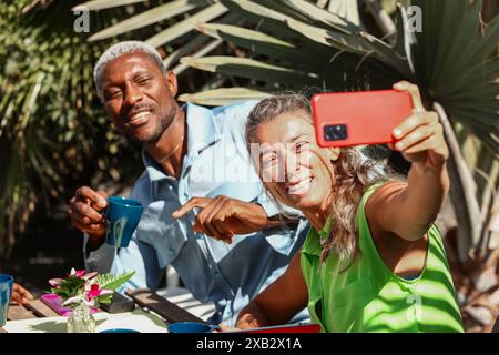 Zwei Freunde genießen ein Sommerfrühstück im Freien, lachen und machen ein Selfie mit der Natur im Hintergrund Stockfoto