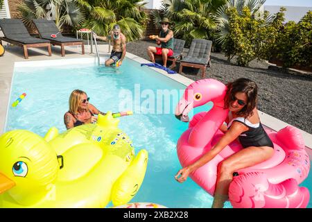 Eine fröhliche Gruppe von Freunden nimmt am Pool Teil, mit farbenfrohen aufblasbaren Schwimmern und verspielten Wasserspielzeugen, im Schatten üppiger Palmen des rel Stockfoto