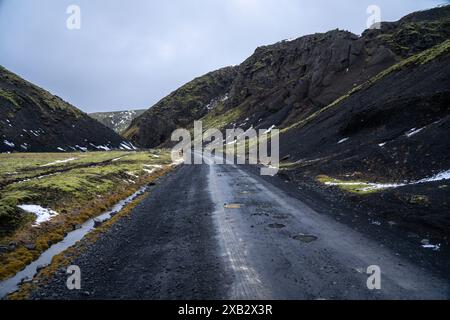 Eine einsame Straße schlängelt sich durch die dramatische Landschaft des Island Highlands, umgeben von steilen Klippen und Schneestücken. Stockfoto